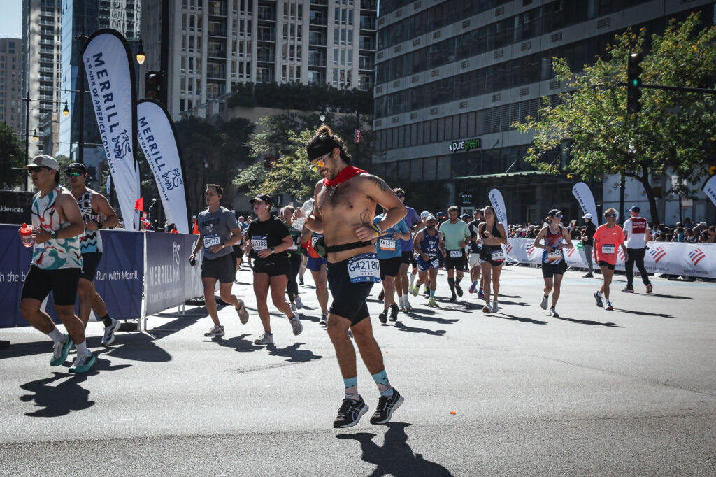 A group of runners participates in the Chicago Marathon on a sunny day in downtown Chicago, with tall buildings, race banners, and spectators lining the street. One runner in the foreground is shirtless, wearing sunglasses, a headband, a red scarf, and themed running socks while mid-stride.