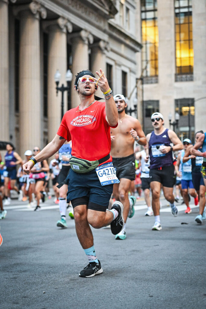 A marathon runner wearing a red Chicago Marathon shirt and race bib G42180 makes a celebratory hand gesture while running through a downtown Chicago street. Other runners follow behind him, with historic stone buildings and glowing windows in the background.